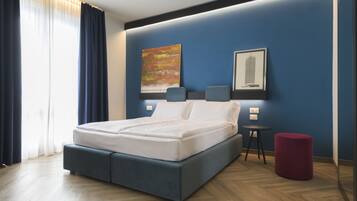 Quarto de hotel moderno com uma cama grande, uma pequena mesa de apoio, um banquinho vermelho e uma pintura com vista para a cidade na parede.