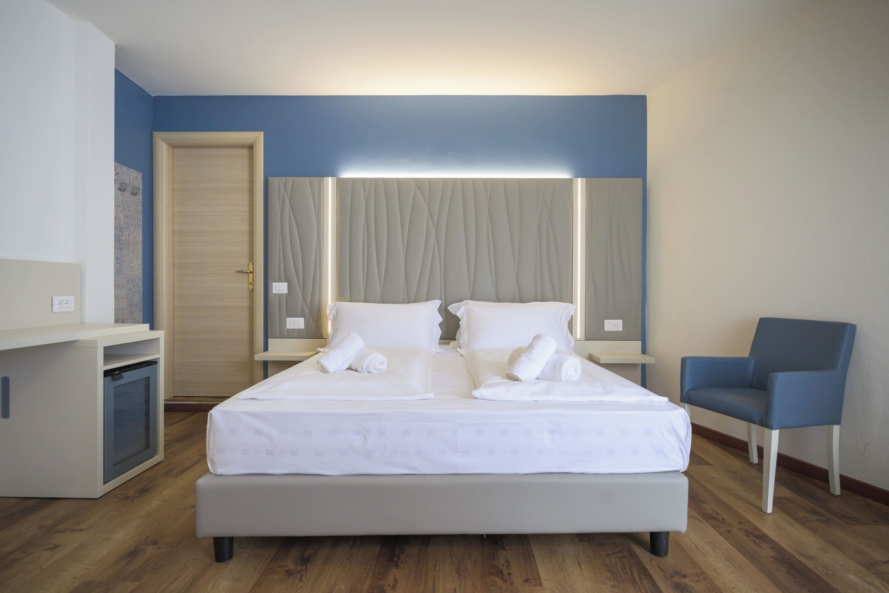 A neatly made double bed with white linens, a bedside table, a chair, and a wall-mounted light fixture.
