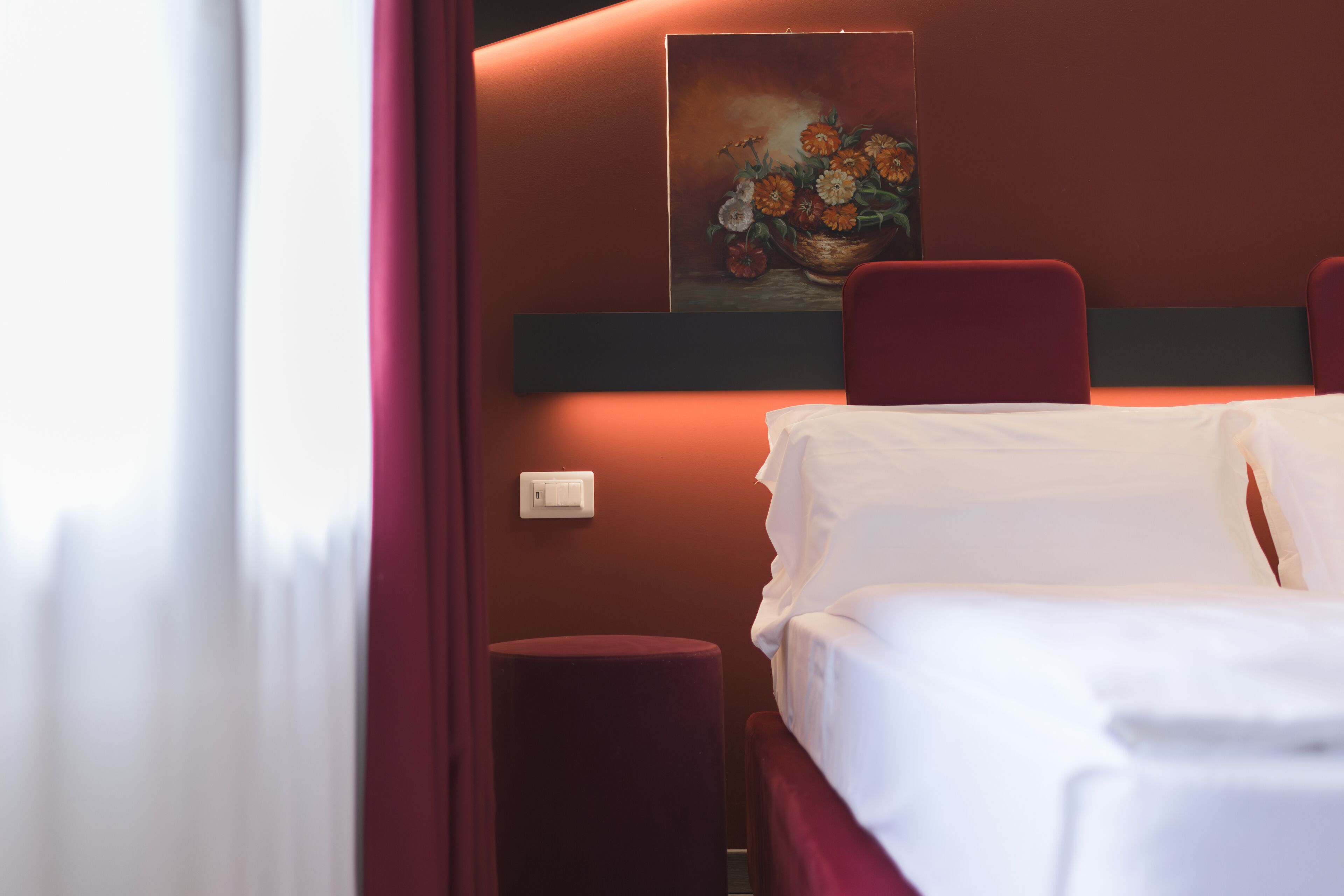 A hotel room with a bed, a red chair, a small table, and a painting on the wall.