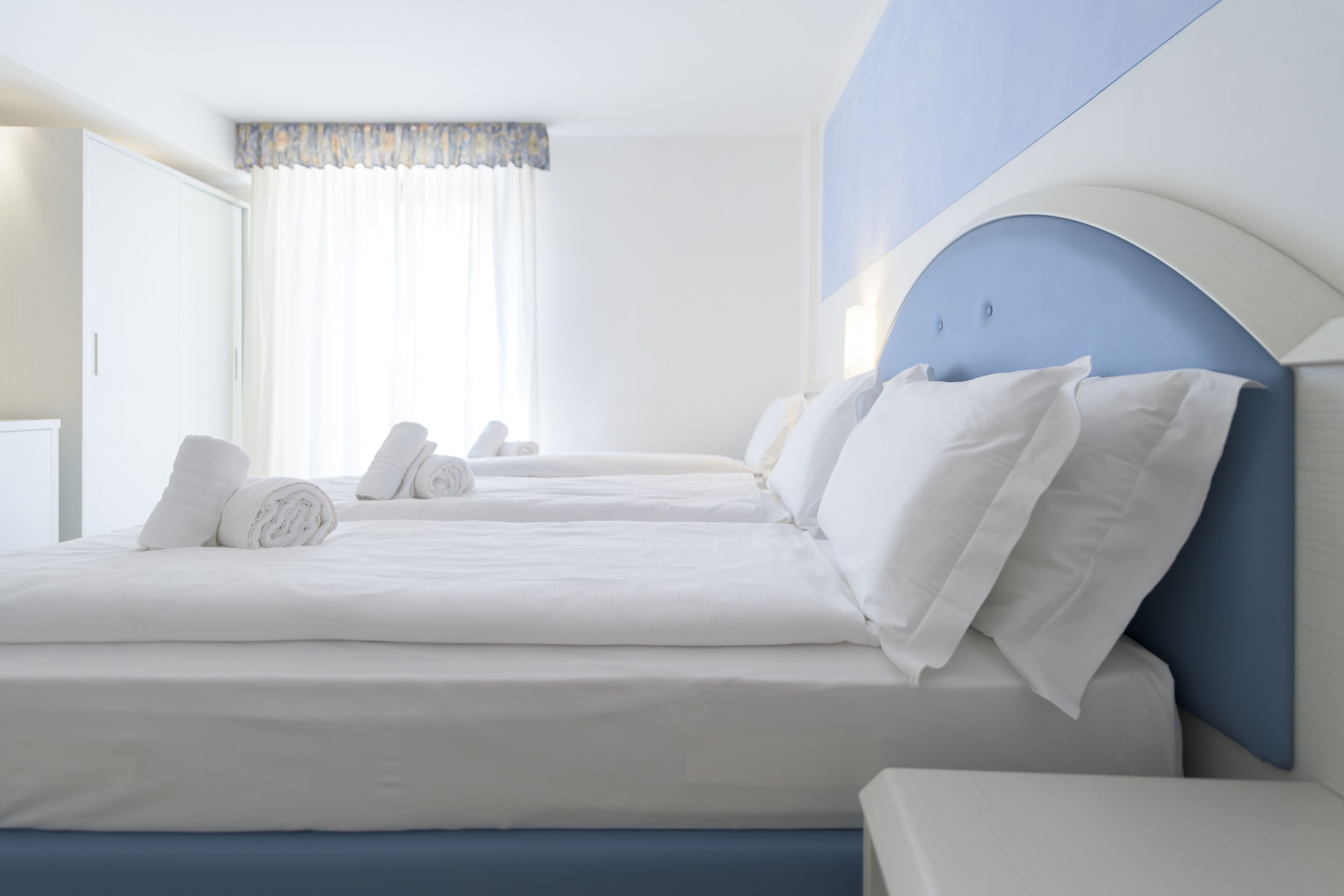 Quarto de hotel com duas camas, roupa de cama branca, uma parede de destaque azul e uma janela com cortinas.