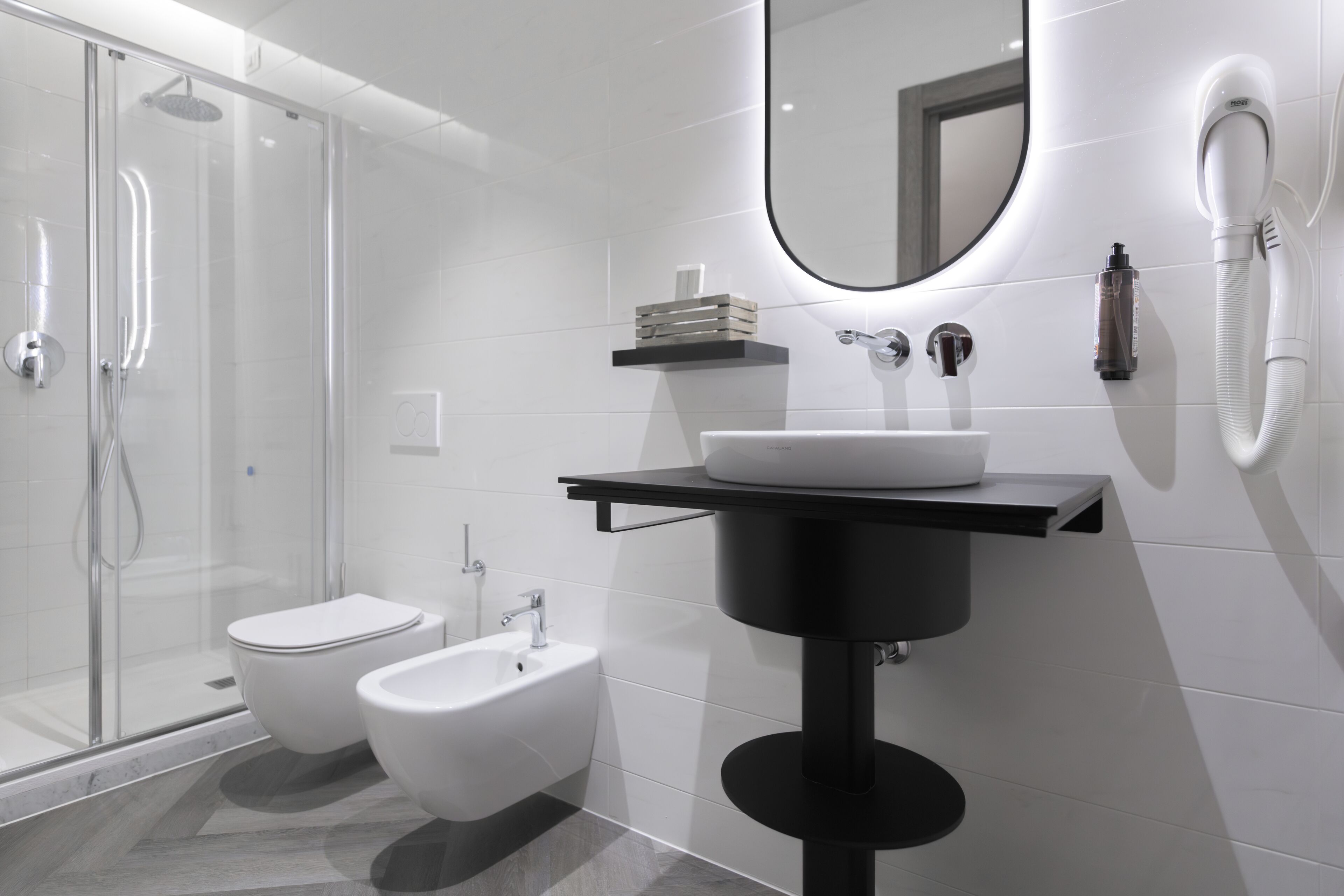 A modern bathroom with a glass-enclosed shower, a bidet, a sink with a black pedestal, and a mirror with a round frame.