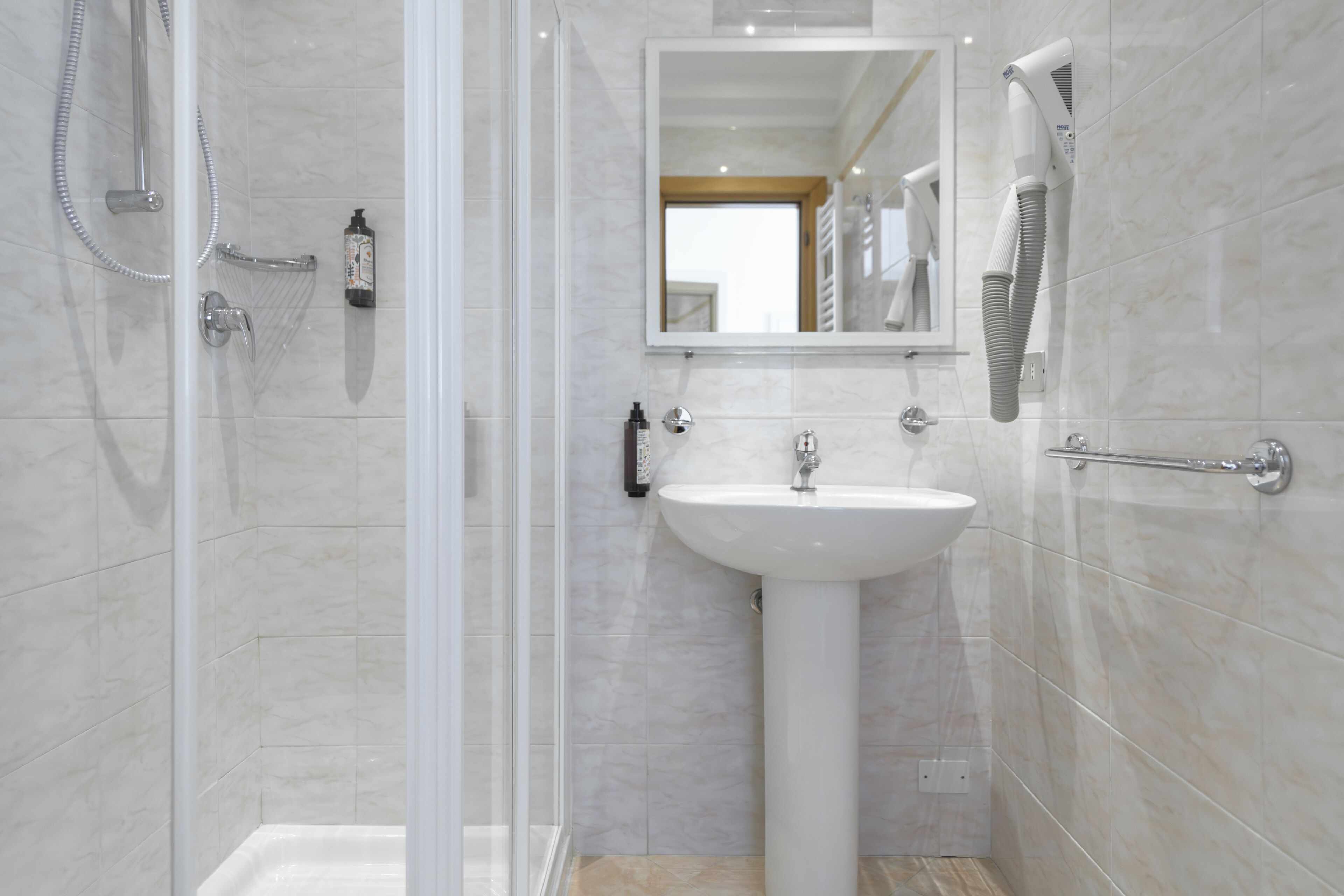 A modern bathroom with a glass-enclosed shower, a pedestal sink, and a handheld hair dryer mounted on the wall.