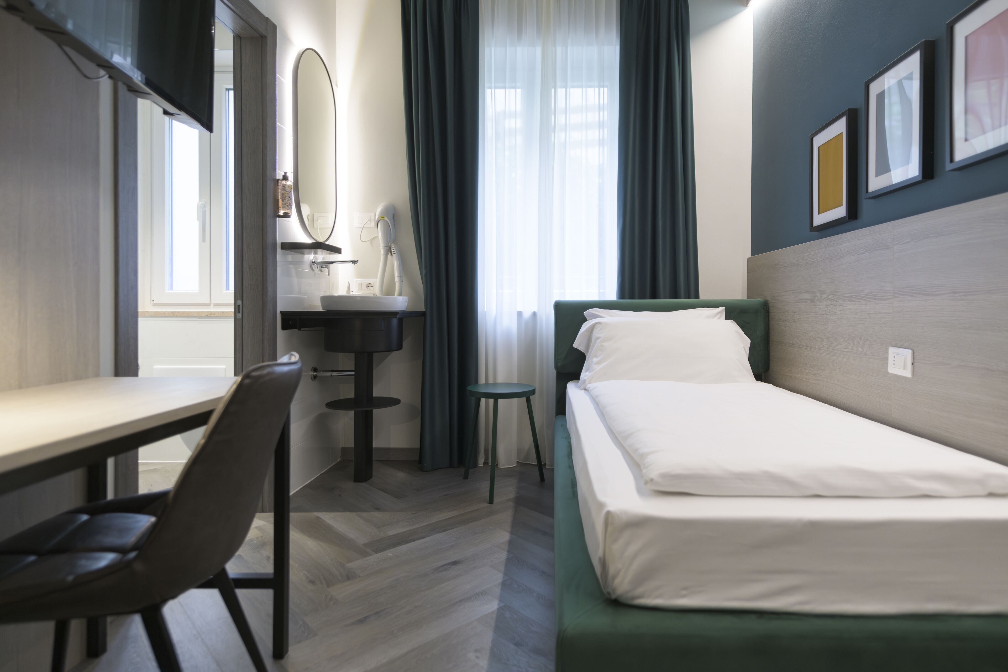 Une chambre d’hôtel équipée d’un lit, d’un bureau, d’une chaise, d’un miroir, d’un lavabo et d’une fenêtre avec des rideaux.