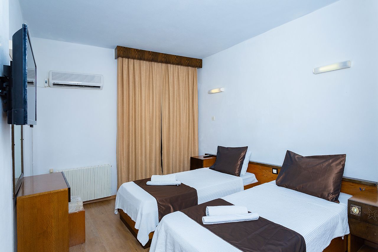 A hotel room with two beds, a TV, an air conditioner, a wooden nightstand, and a window with curtains.