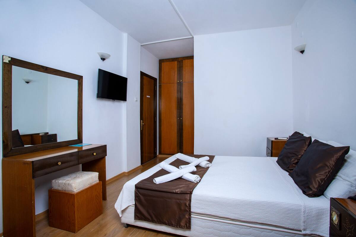 A bedroom with a bed, a wooden desk, a mirror, a television, and a wardrobe.