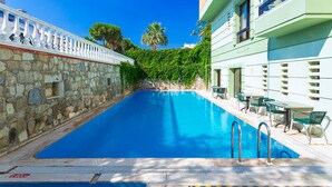 Una piscina con paredes de piedra y balaustrada blanca, rodeada de vegetación y un edificio con balcones.