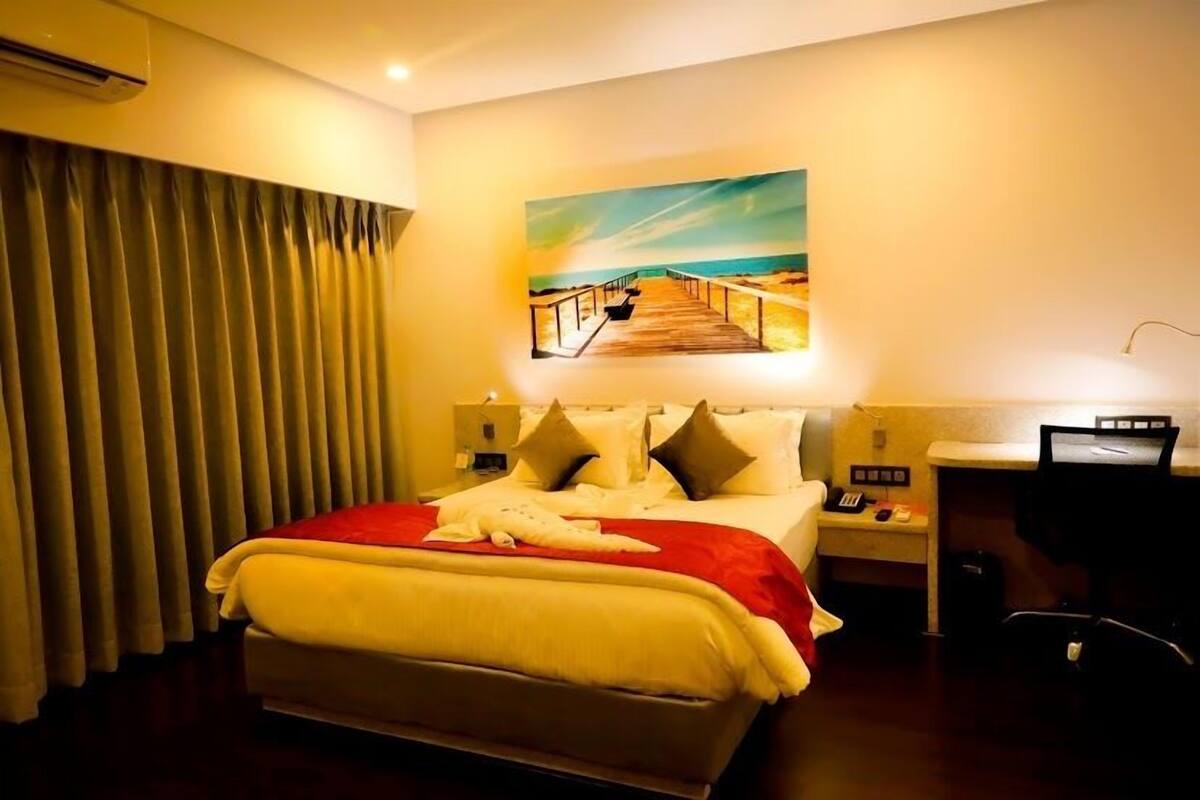 A hotel room with a bed, a painting of a beach, a desk with a computer, and a nightstand with a phone.