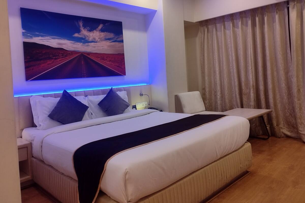 A modern hotel room with a large bed, a bedside table, a chair, a wall-mounted air conditioner, and a large landscape painting above the bed.