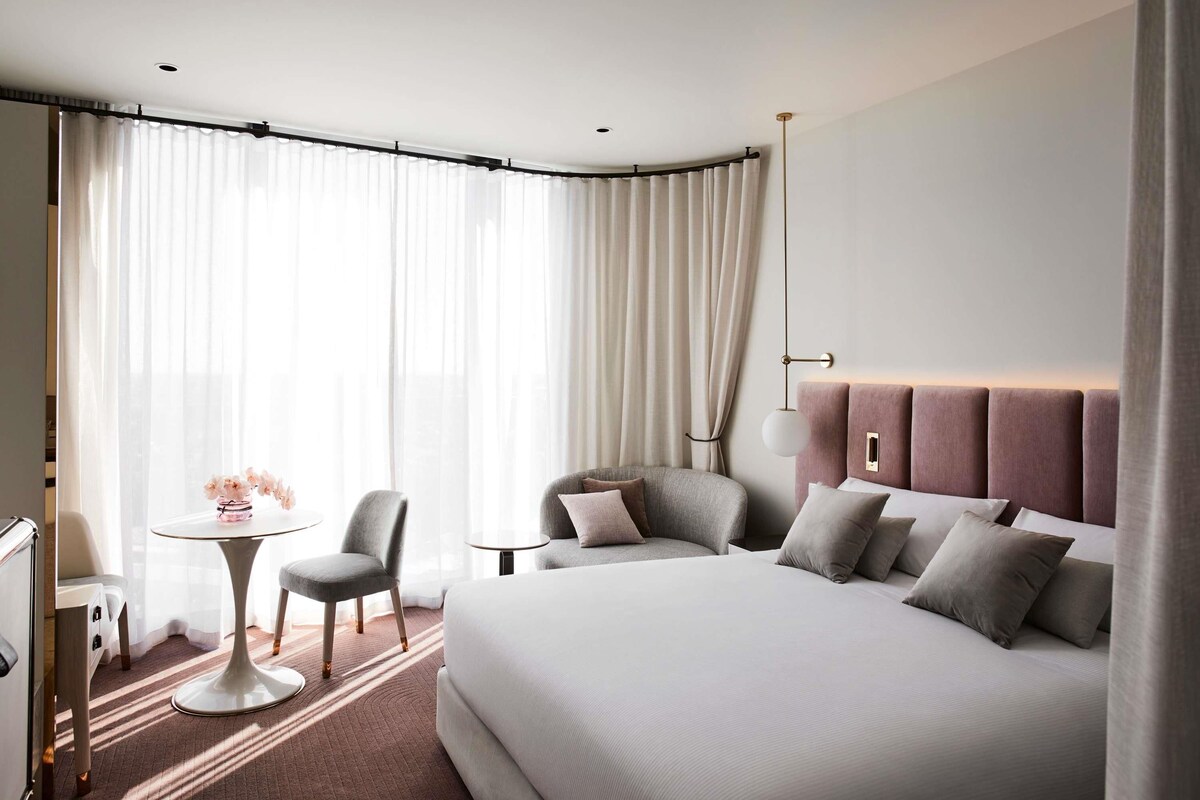 A modern hotel room with a large bed, a desk, a chair, and a small table.