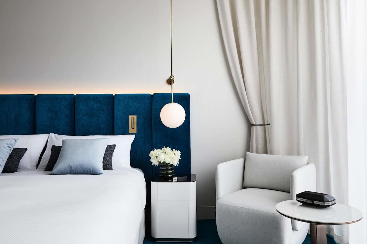 A modern hotel room with a large bed, a bedside table with a vase of flowers, a round table with a phone, and a chair.