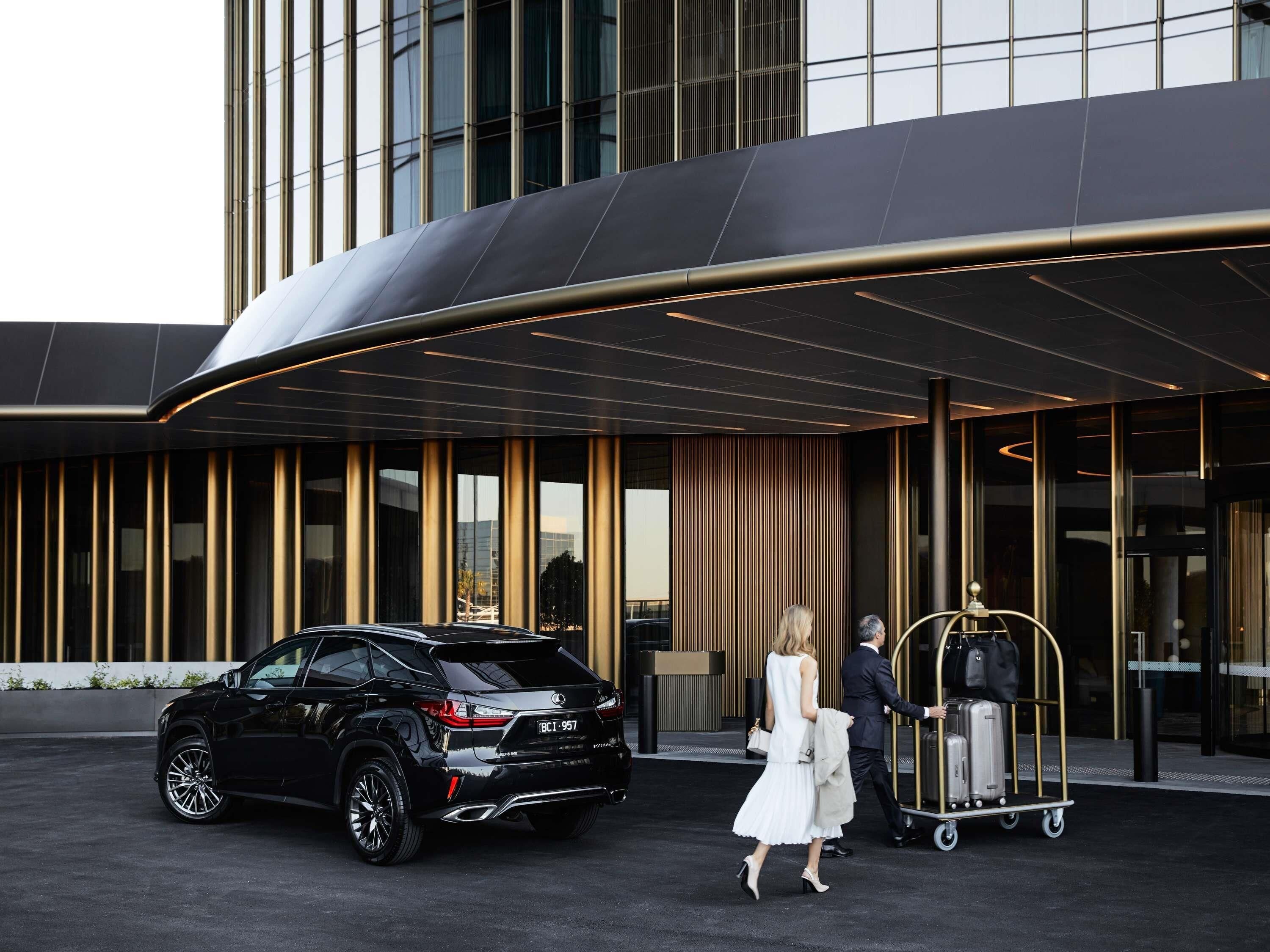 A modern building with a curved entrance, a black SUV parked in front, and a luggage cart with suitcases.