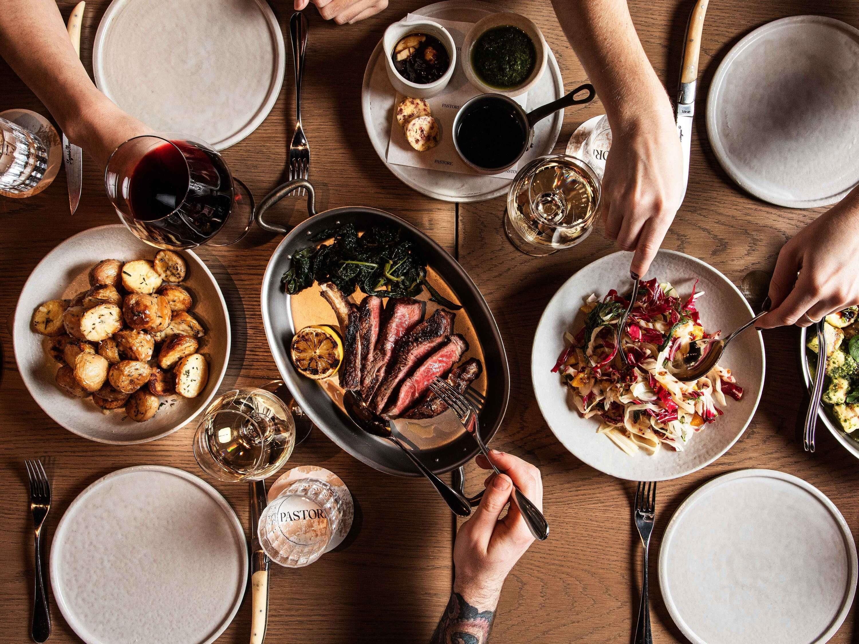A dining table with various dishes, including roasted potatoes, a steak, and a salad, along with multiple glasses of wine.