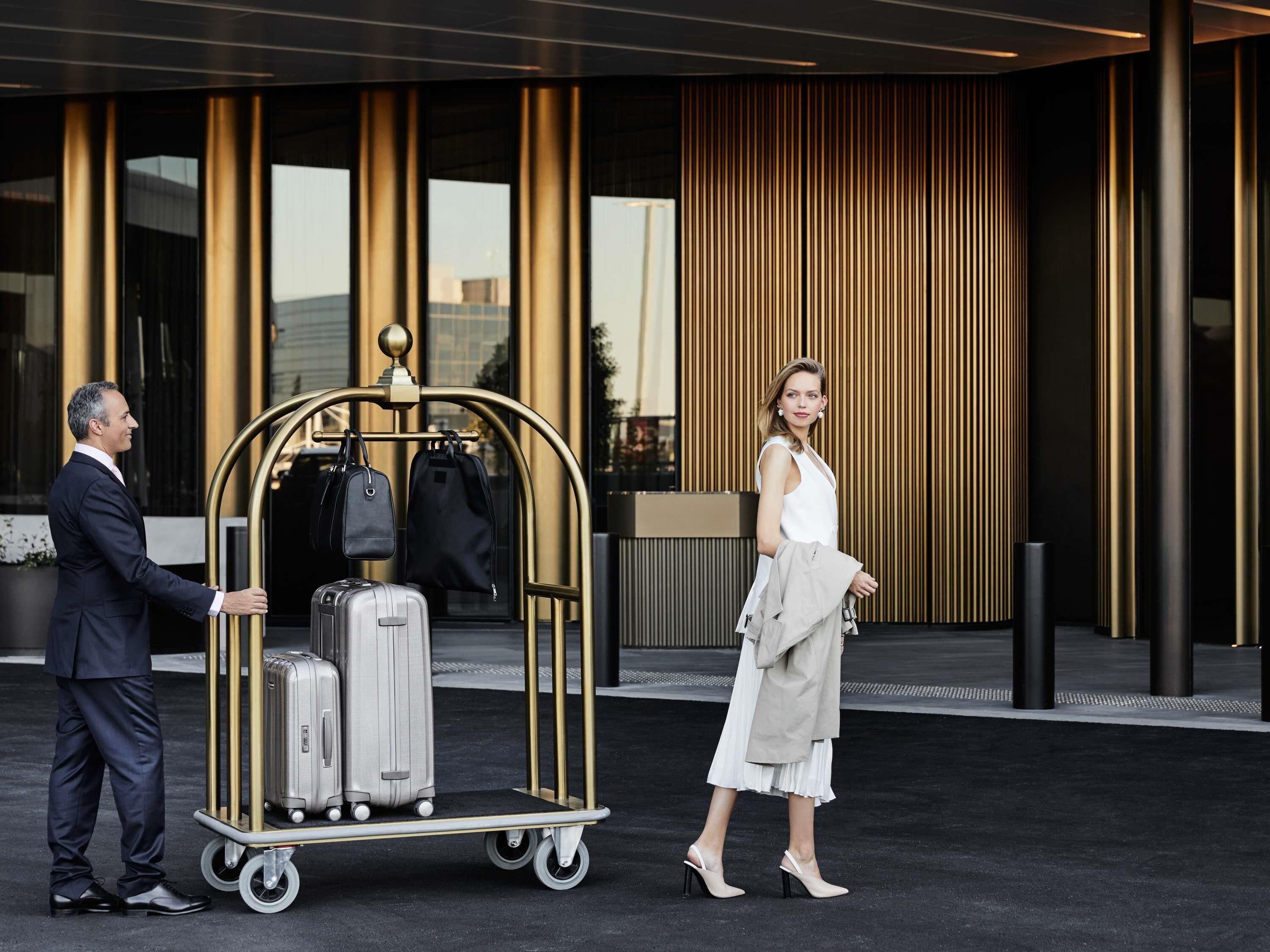 Un homme poussant un chariot à bagages rempli de valises et de sacs, une femme en robe blanche, et un bâtiment aux lignes verticales et à la façade dorée.