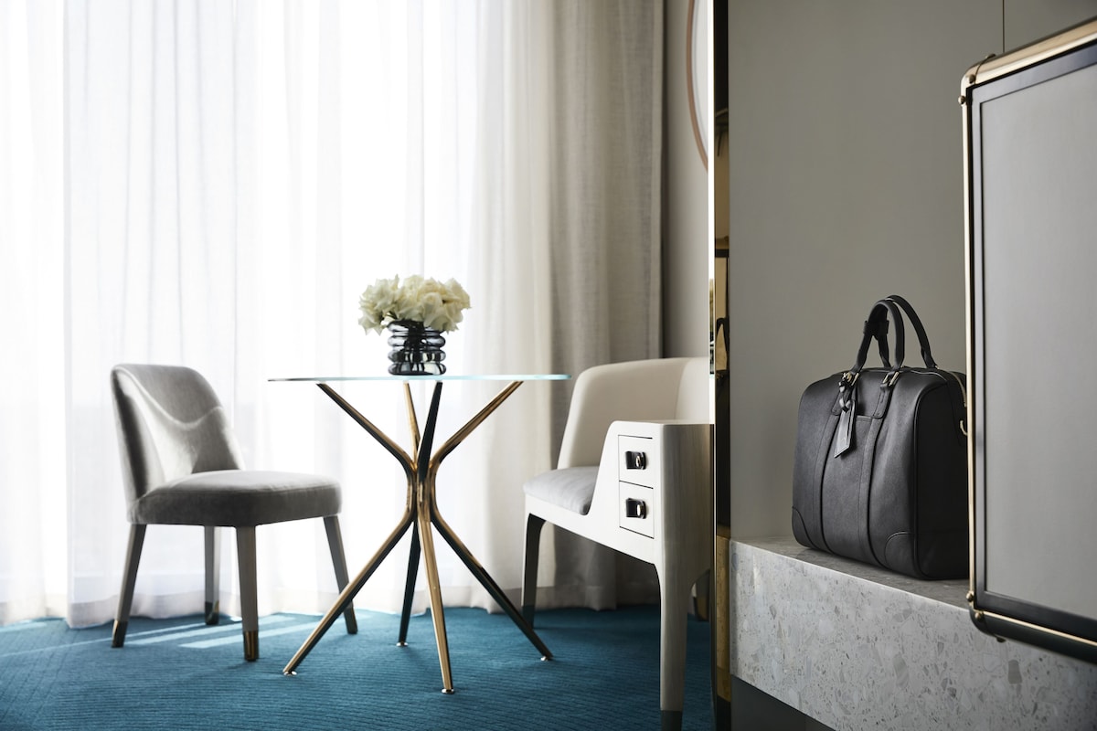 Et hotelværelse med et glasbord, to stole, en vase med blomster, en sort kuffert og et stort spejl.