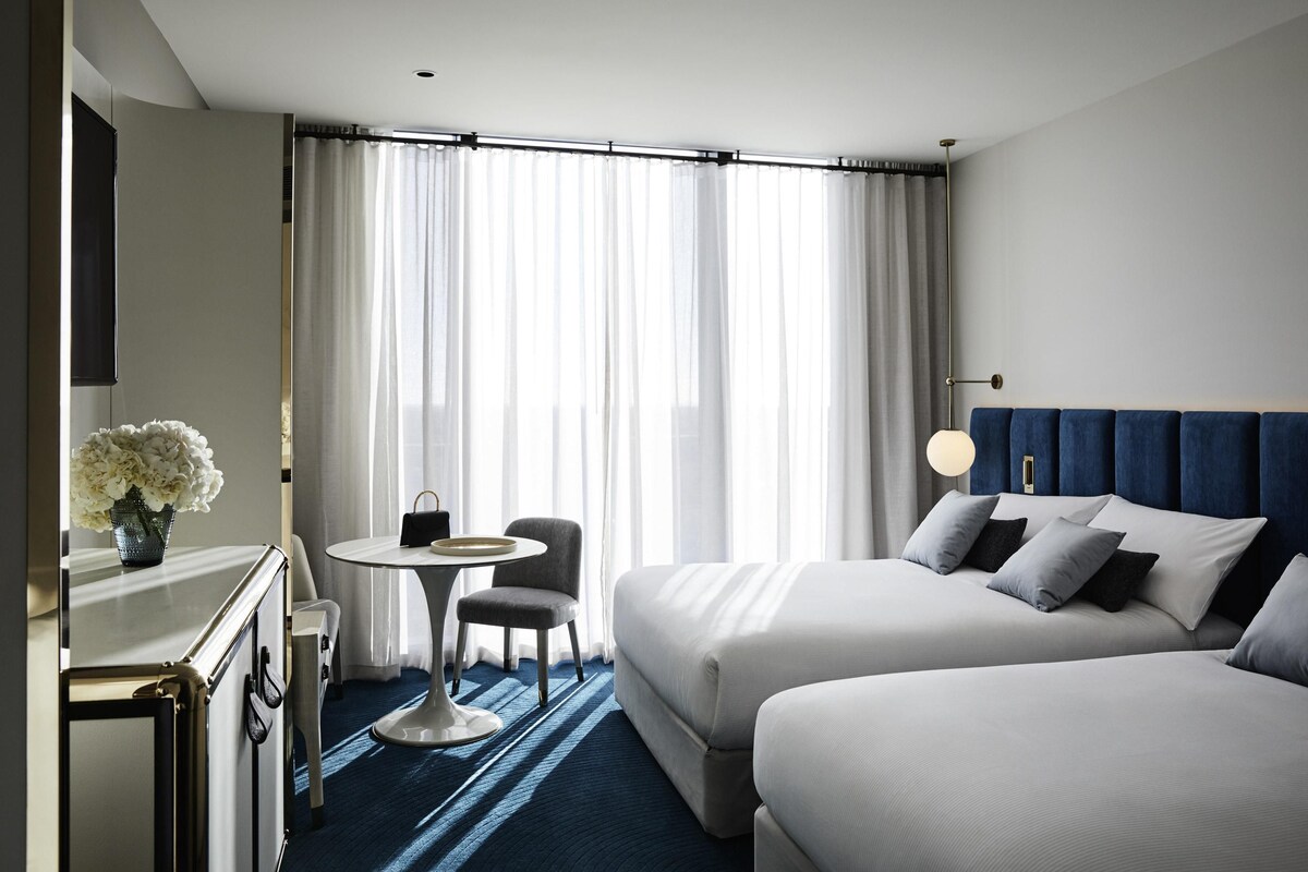 A hotel room with two beds, a small table with a chair, a vase with flowers, and a large window with curtains.