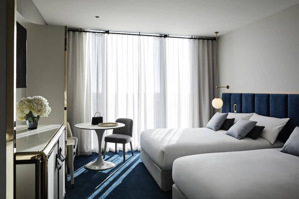 A hotel room with two beds, a small table with a chair, a vase with flowers, and a large window with curtains.