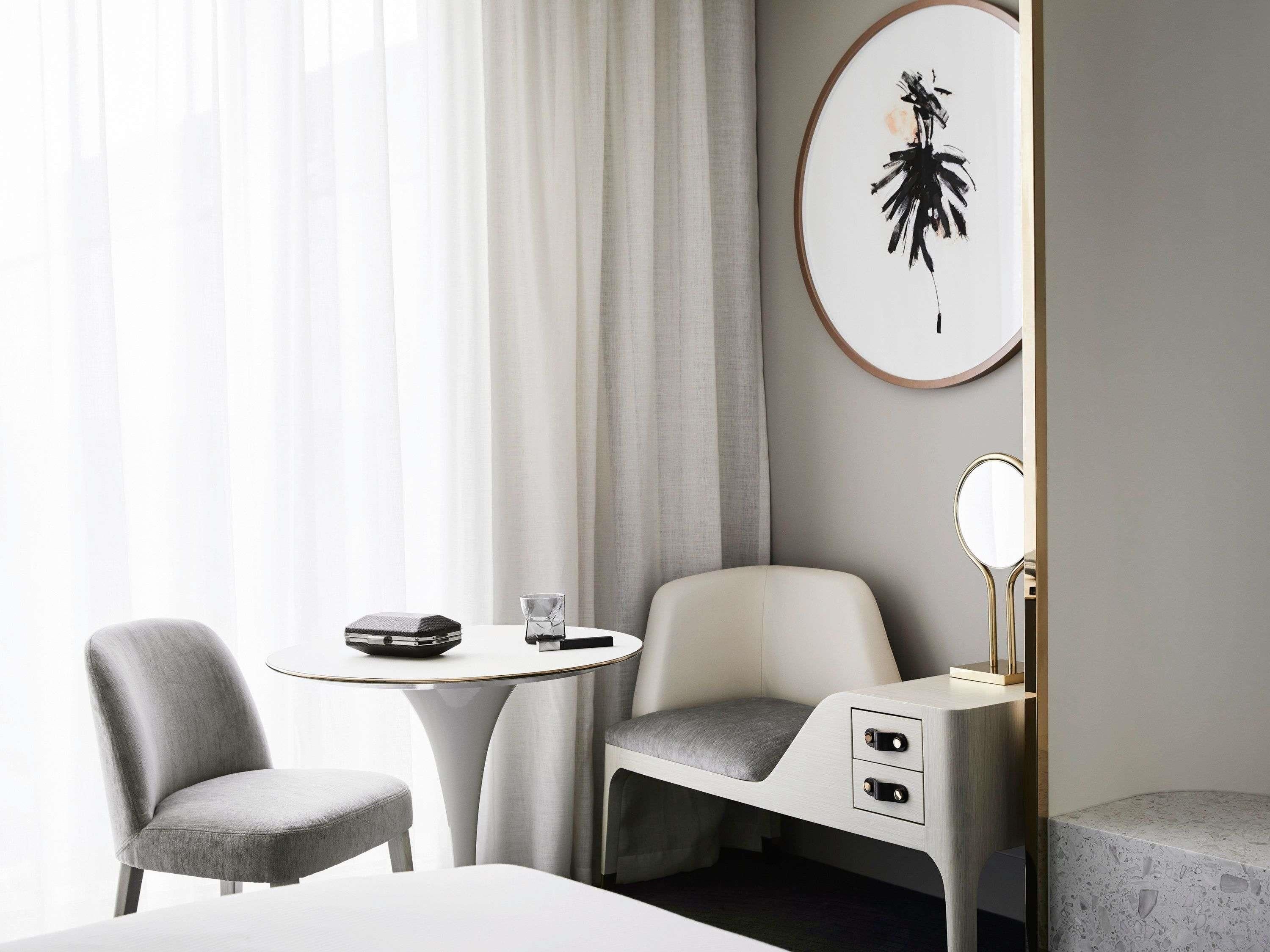 A modern hotel room with a bed, a desk with a chair, a small table, and a wall-mounted mirror.