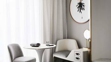 A modern hotel room with a bed, a desk with a chair, a small table, and a wall-mounted mirror.