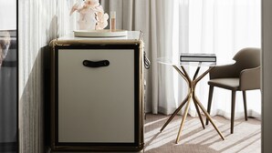 Una habitación de hotel moderna con diseño minimalista, que incluye una mesita de noche con un jarrón de flores, un espejo redondo y una original mesita auxiliar de patas múltiples.