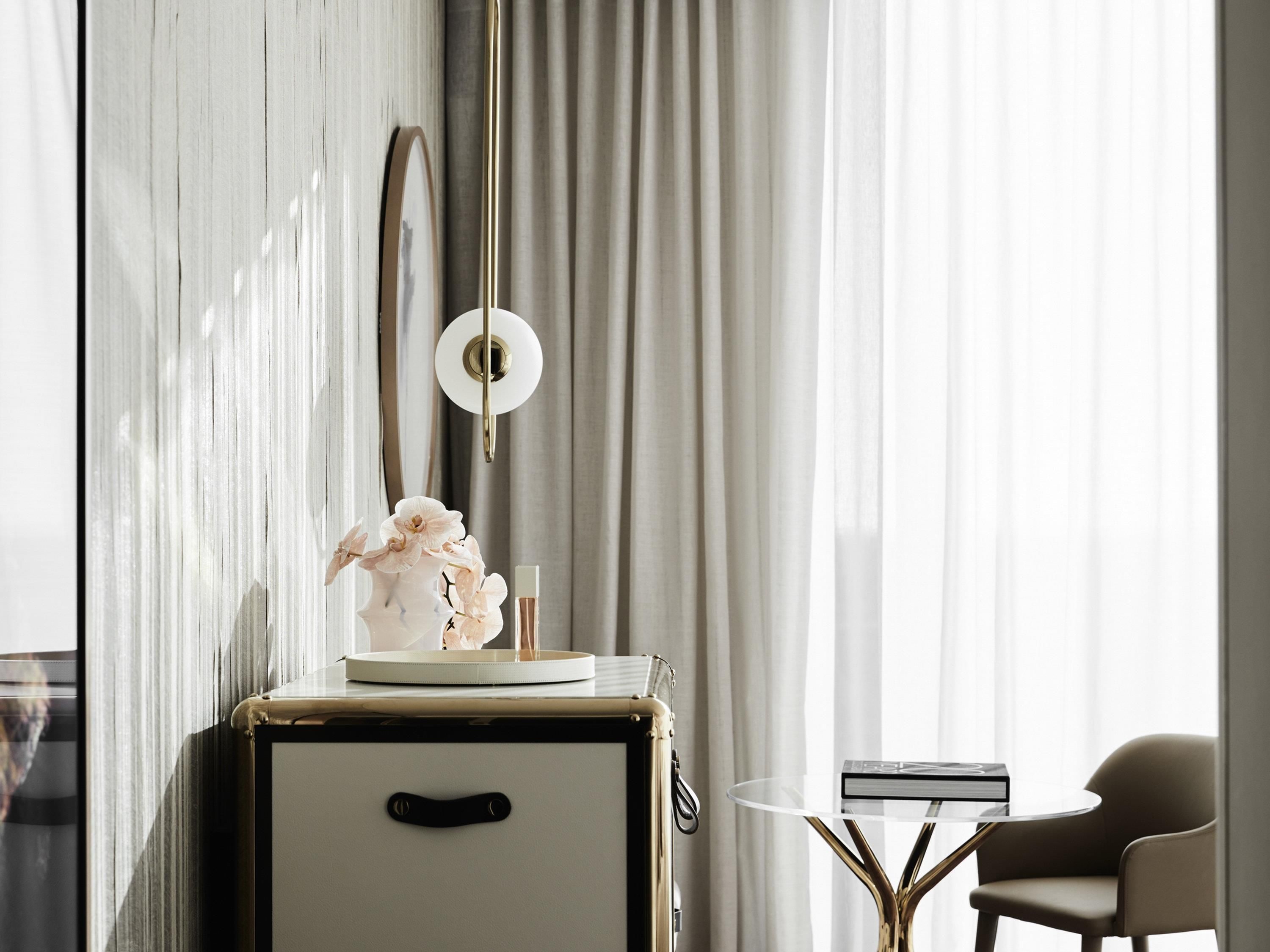 A room with a vintage suitcase, a small table with a book, a chair, a wall-mounted light fixture, and a framed picture.