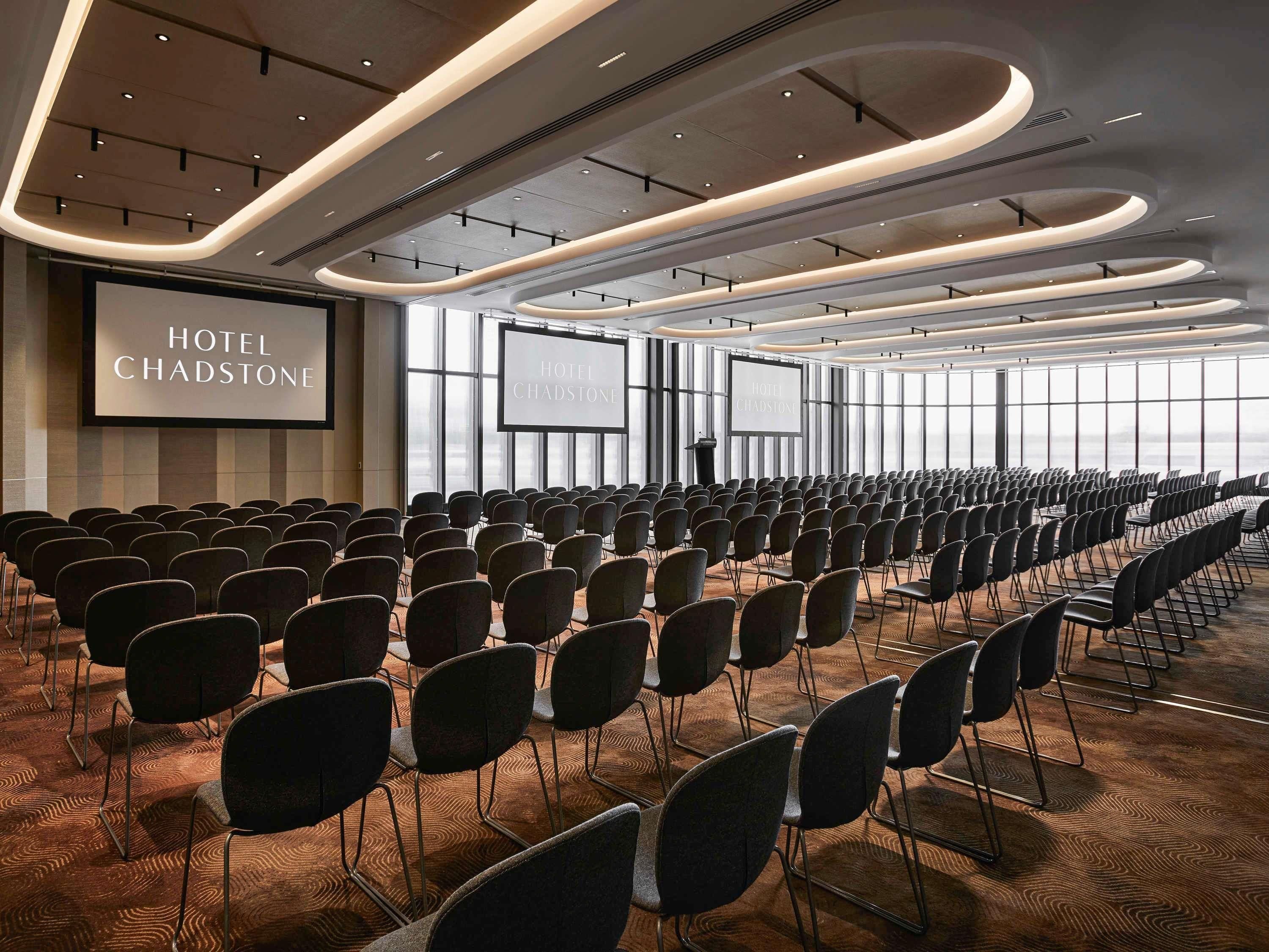 Une salle de conférence avec des rangées de chaises, de grands écrans affichant le nom « HÔTEL CHADSTONE » et un pupitre.