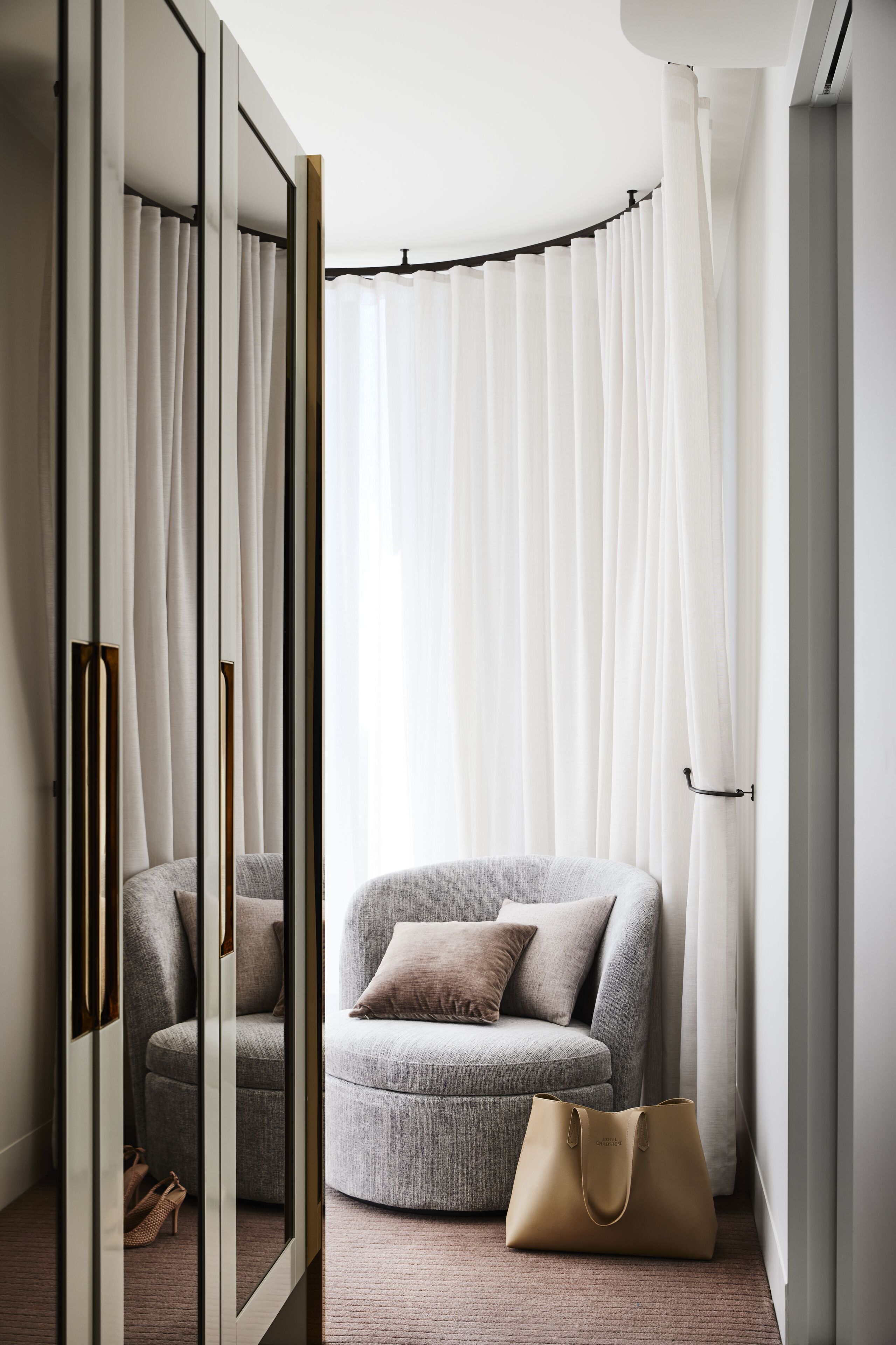 Une pièce moderne avec un canapé gris, un sac à main beige et un miroir qui reflète la pièce.