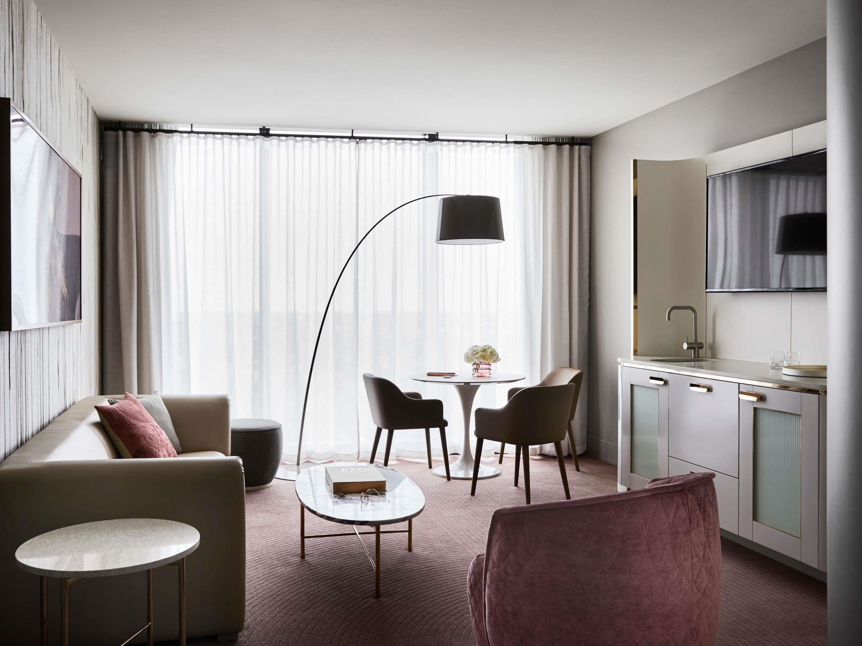 A modern hotel room with a sofa, a small round table, a dining area with chairs, a kitchen area, and a large window with curtains.