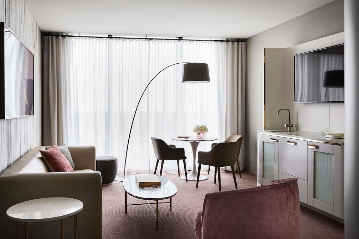 A modern hotel room with a sofa, a small round table, a dining area with chairs, a kitchen area, and a large window with curtains.