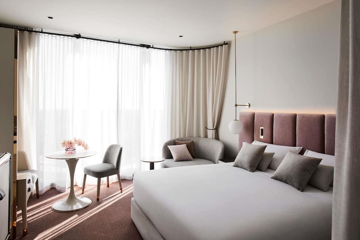 A modern hotel room with a large bed, a desk, a chair, and a small table.