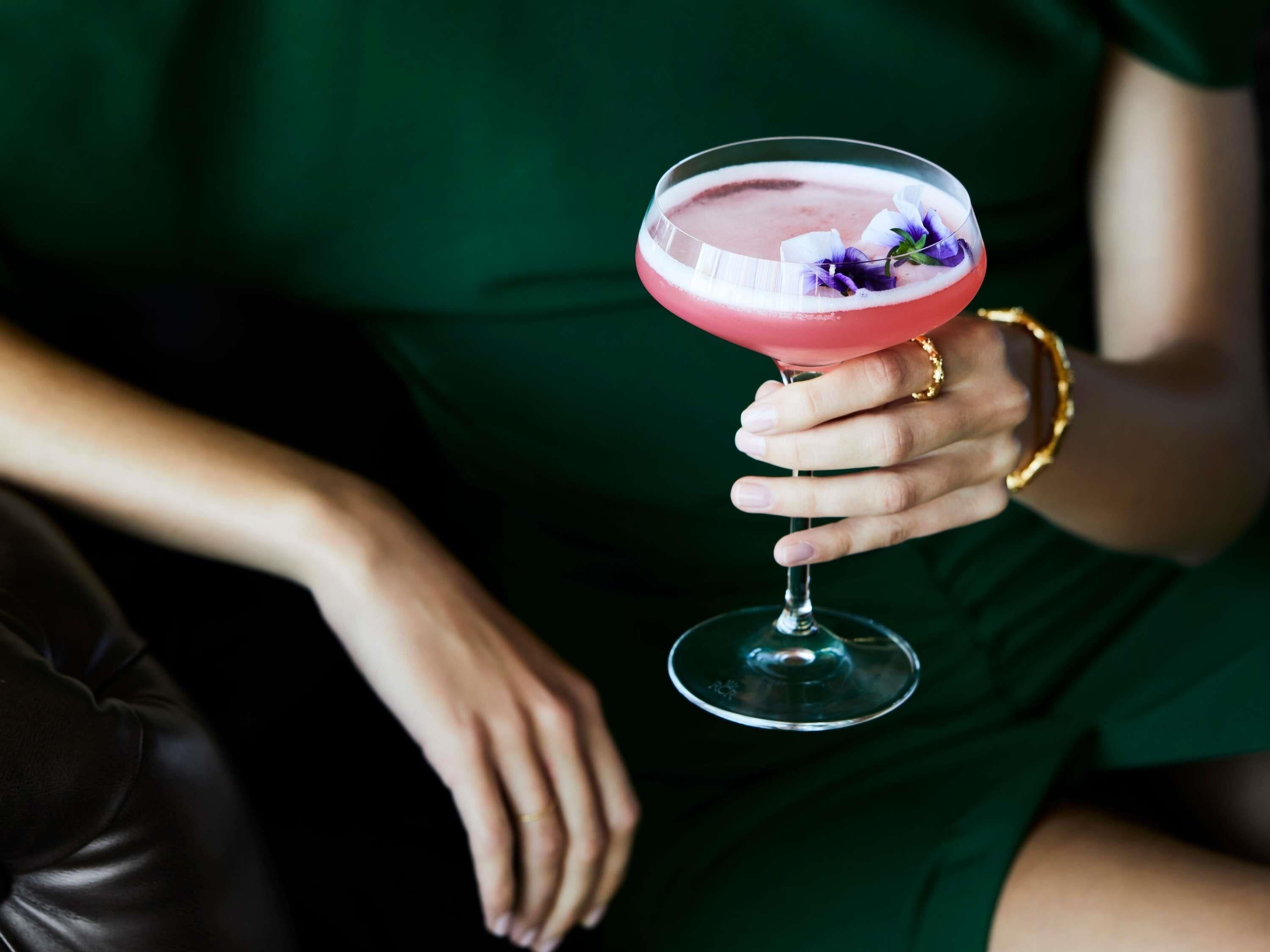 A person holding a pink cocktail with a flower garnish.