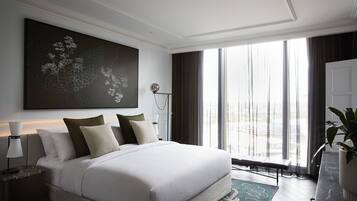 A modern hotel bedroom with a large bed, a nightstand, a lamp, a large window with curtains, and a painting on the wall.