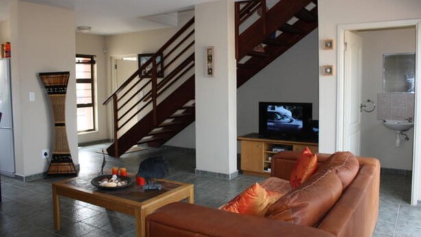 A living room with a sofa, a coffee table, a television, a staircase, and a kitchen area. - On The Dunes 8 (Henties Bay)