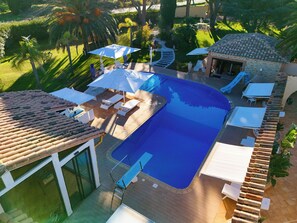 Une piscine entourée d’un jardin et d’un bâtiment à toit de tuiles, avec des chaises longues et des parasols à proximité.