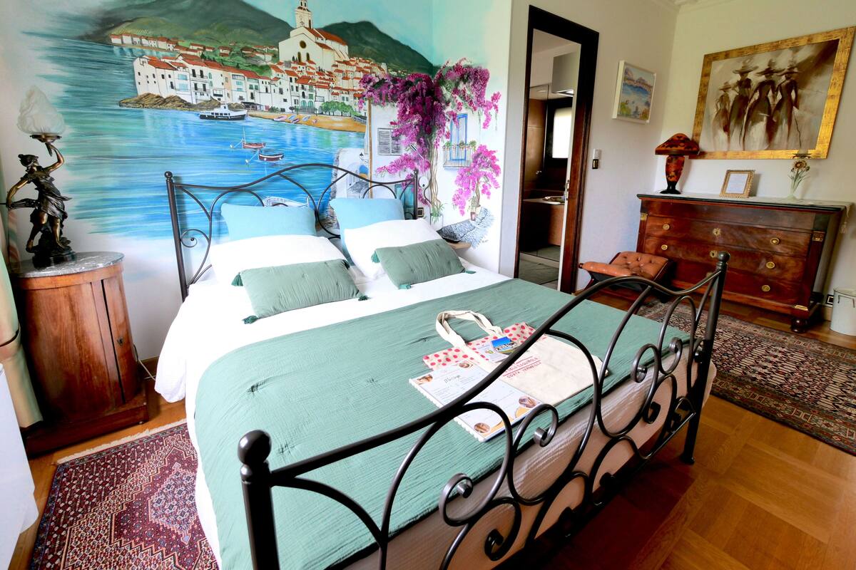 Ein Schlafzimmer mit Bett, Schminktisch, Nachttisch, einem Bild einer Küstenstadt und einem Sessel.