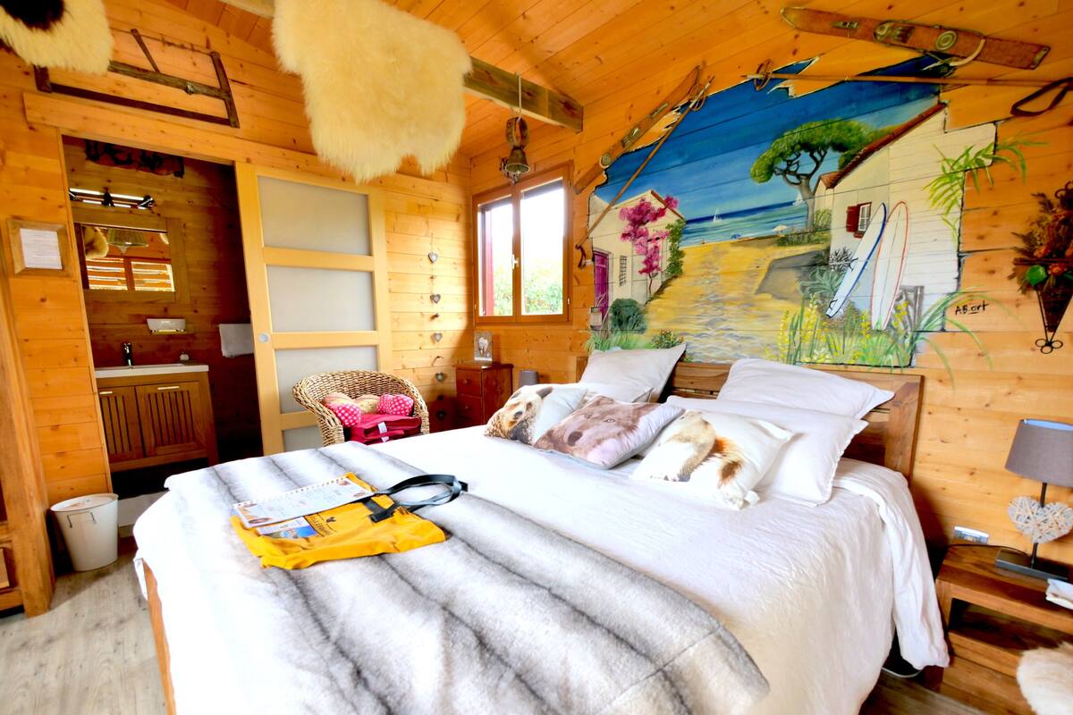 Ein gemütliches Holzblockhaus-Zimmer mit einem Bett, einem Korbstuhl und einem Wandgemälde.