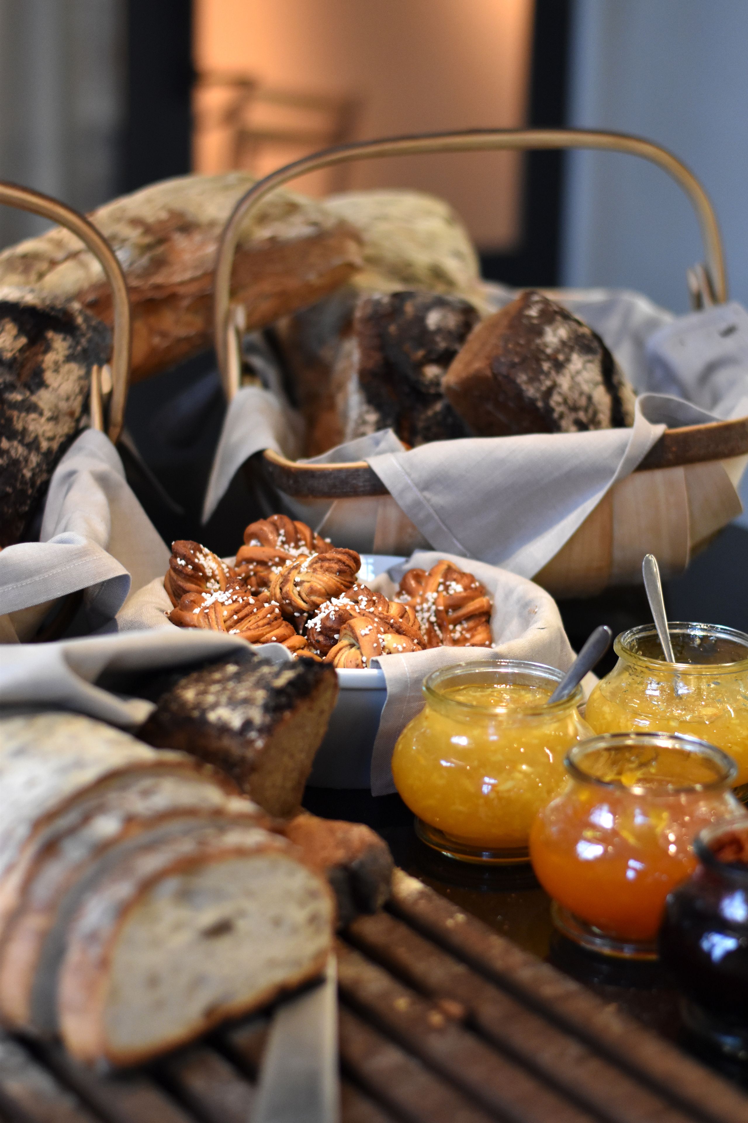Une variété de pains et de viennoiseries disposés sur une table en bois, accompagnés de deux pots de confiture d’orange.