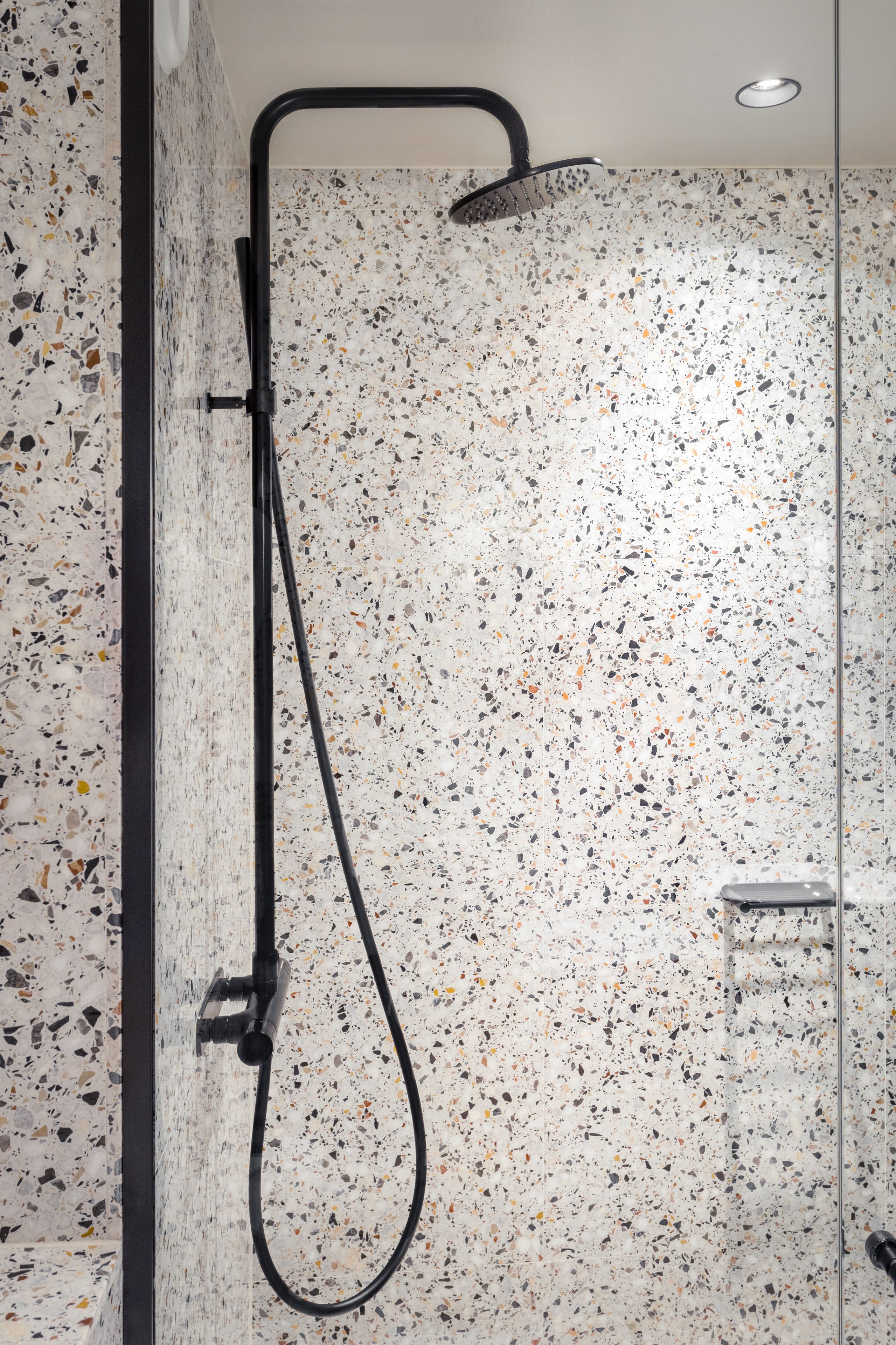 A shower with a black hose and a handheld showerhead, set against a speckled tile wall.