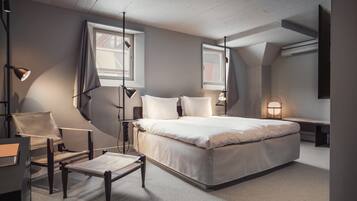 Ein modernes Schlafzimmer mit einem großen Bett, einem Schreibtisch und einem Stuhl. Zwei Fenster sorgen für natürliches Licht, und ein Flachbildfernseher ist an der Wand montiert.