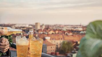 A person pouring a drink into a glass, with a bottle and another glass in the foreground, and a cityscape in the background.