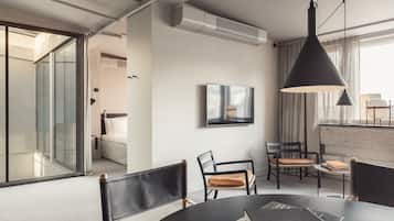 Una habitación de hotel moderna con comedor, sofá, televisor y un amplio ventanal con cortinas.