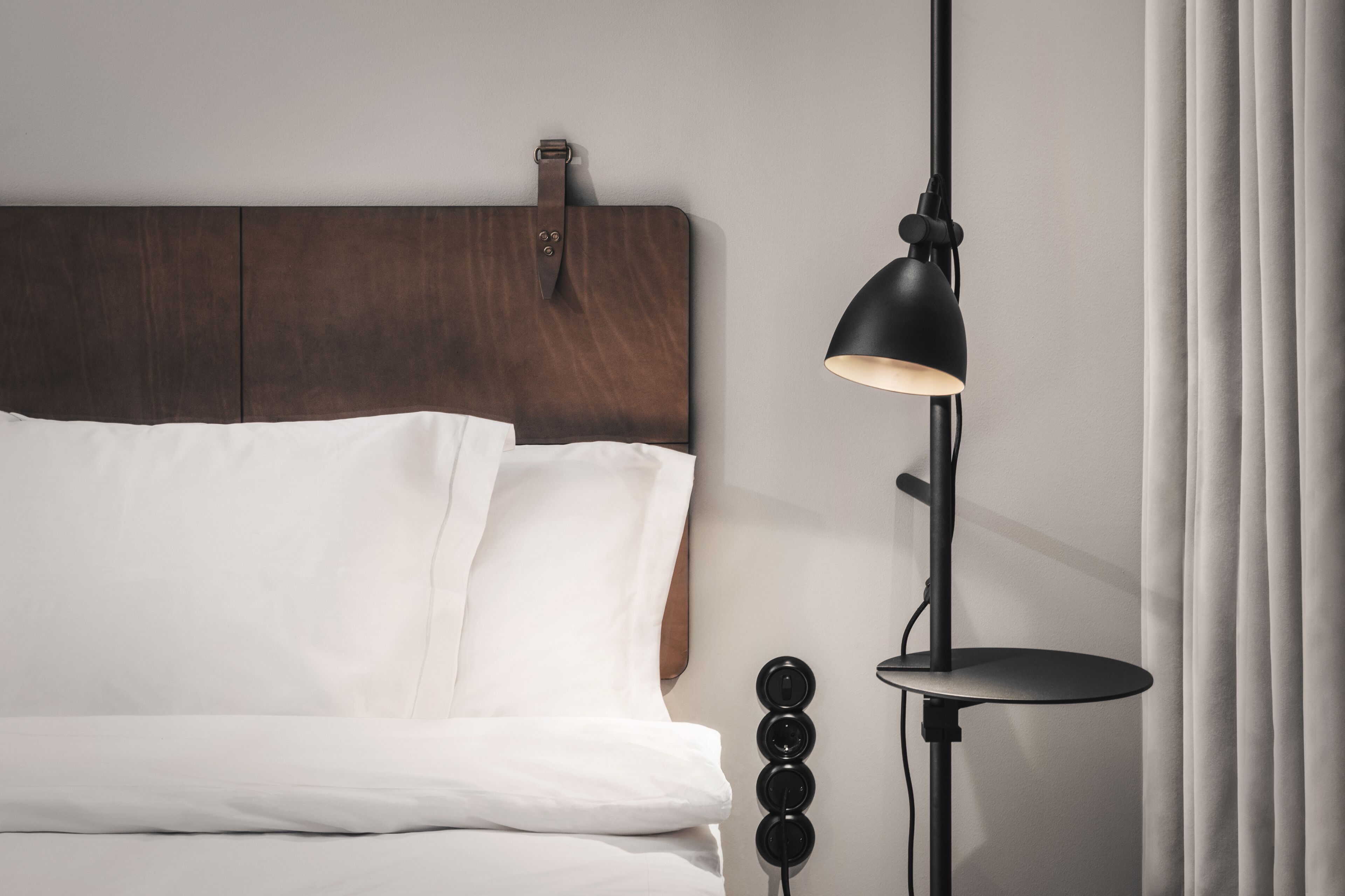 Una cama bien tendida con sábanas blancas, un cabecero de madera oscura y una lámpara de pie negra con una mesita redonda al lado.