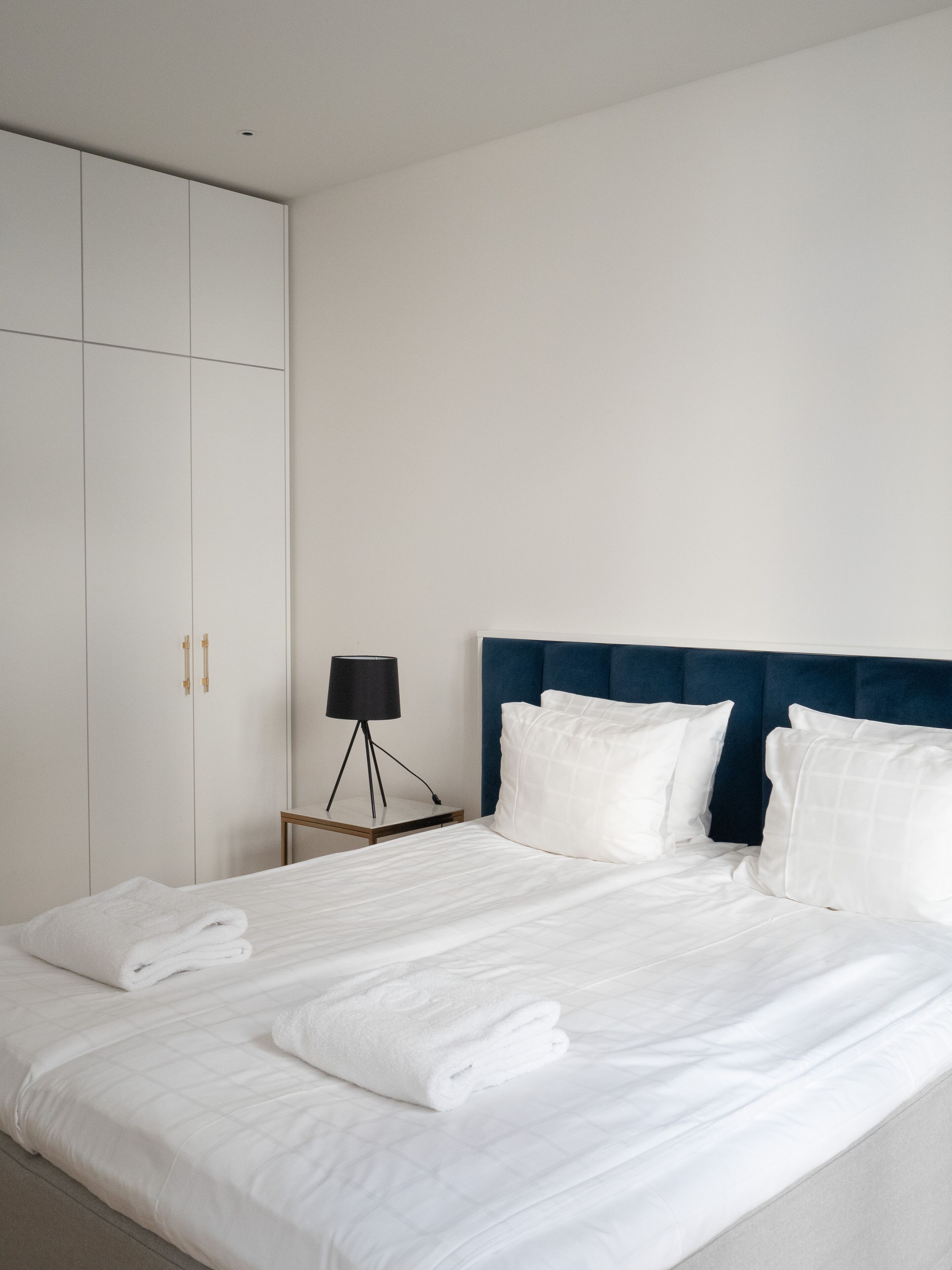 Una cama bien hecha con sábanas y toallas blancas, un cabecero azul oscuro y una mesita de noche con una lámpara.