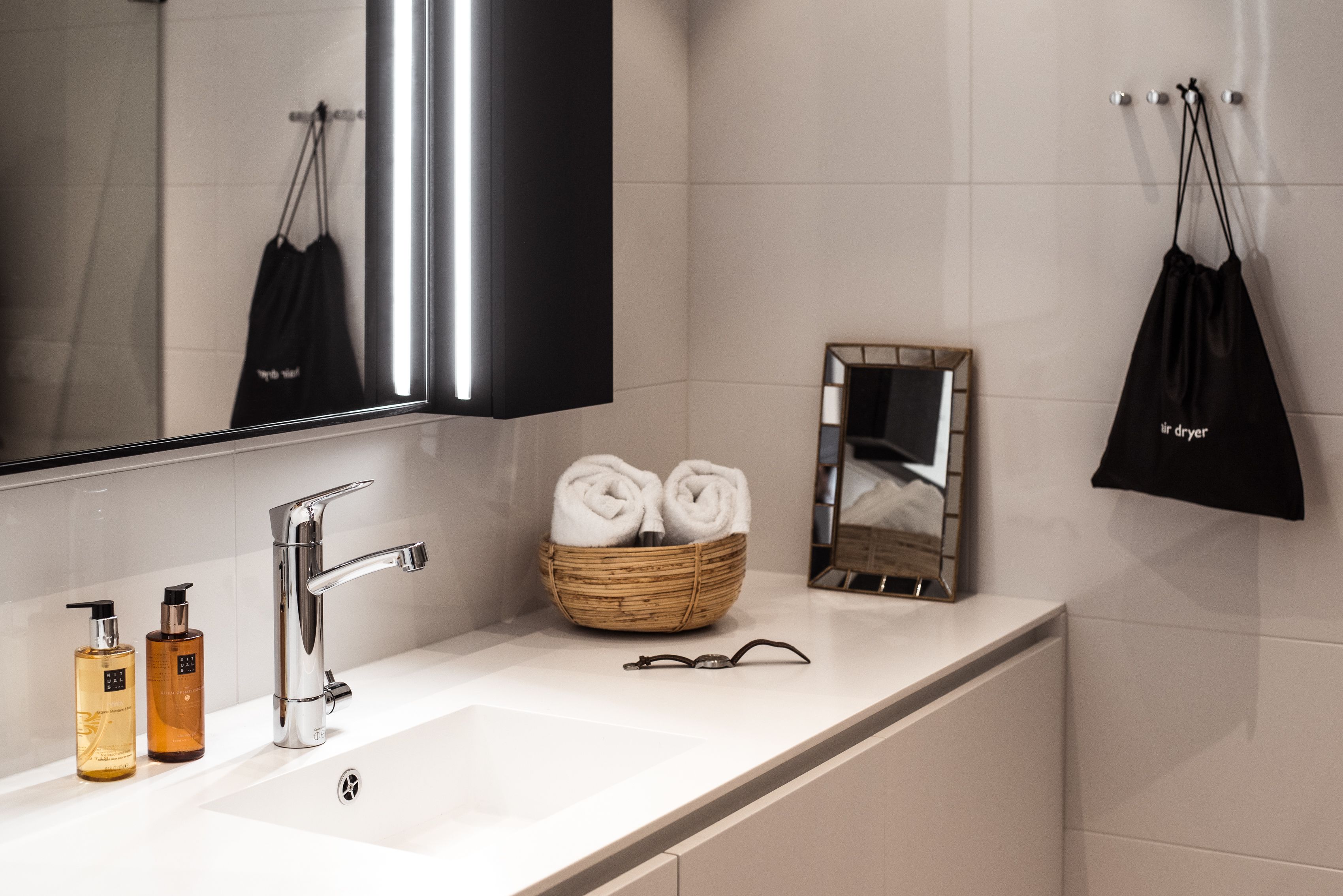 A modern bathroom with a large mirror, a chrome faucet, a white sink, and a wicker basket holding rolled towels.