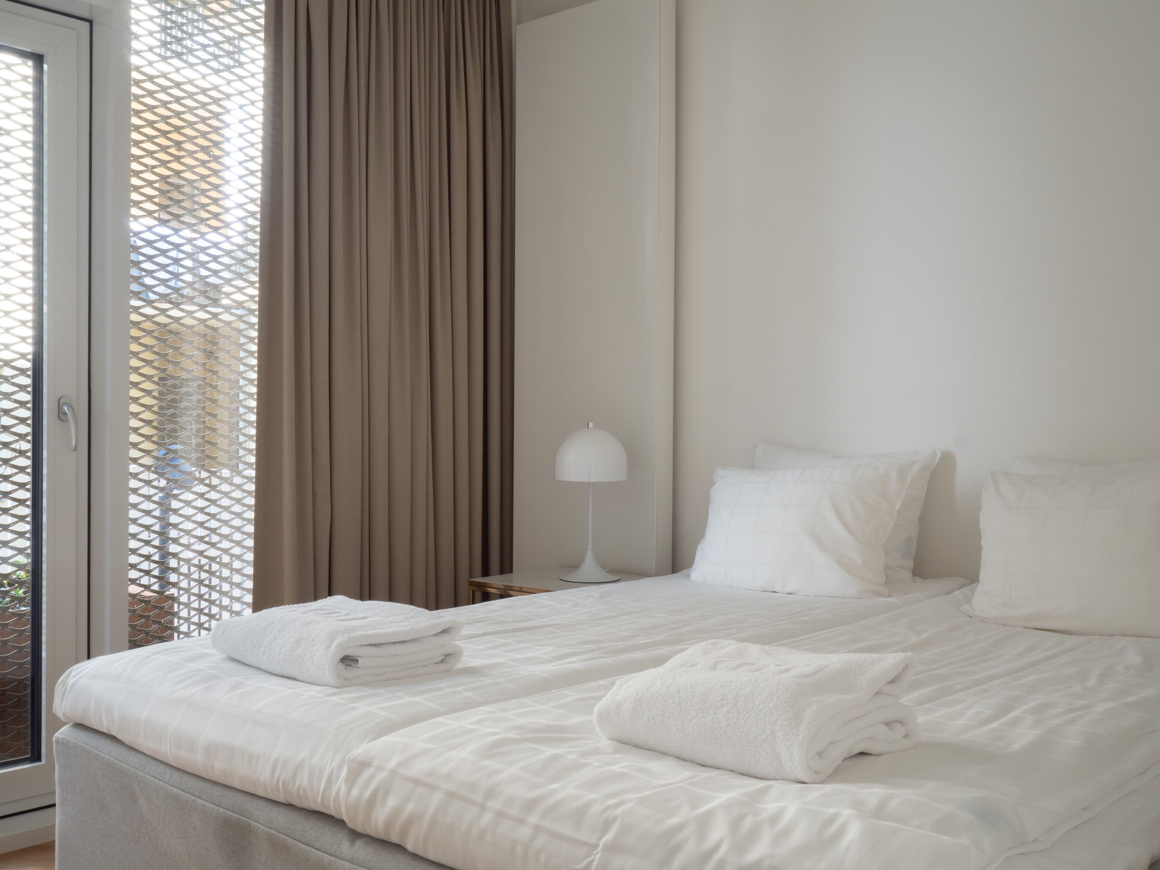 Une chambre d’hôtel avec un lit, du linge de lit blanc, une lampe et une vue sur l’extérieur à travers une fenêtre avec des rideaux.
