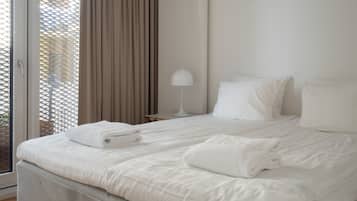 Une chambre d’hôtel avec un lit, du linge de lit blanc, une lampe et une vue sur l’extérieur à travers une fenêtre avec des rideaux.