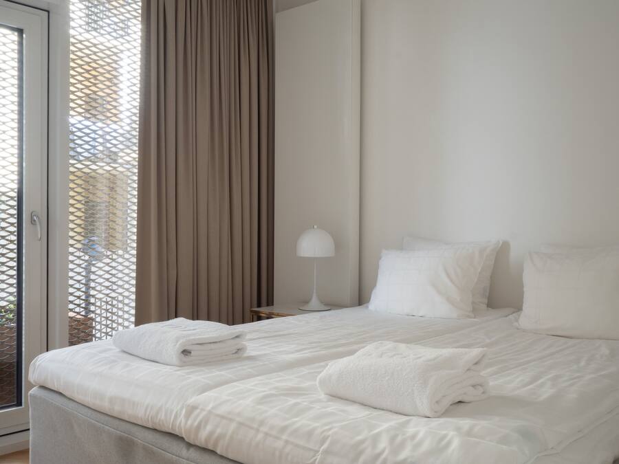 Habitación de hotel con cama, ropa de cama blanca, lámpara y vista a través de una ventana con cortinas.