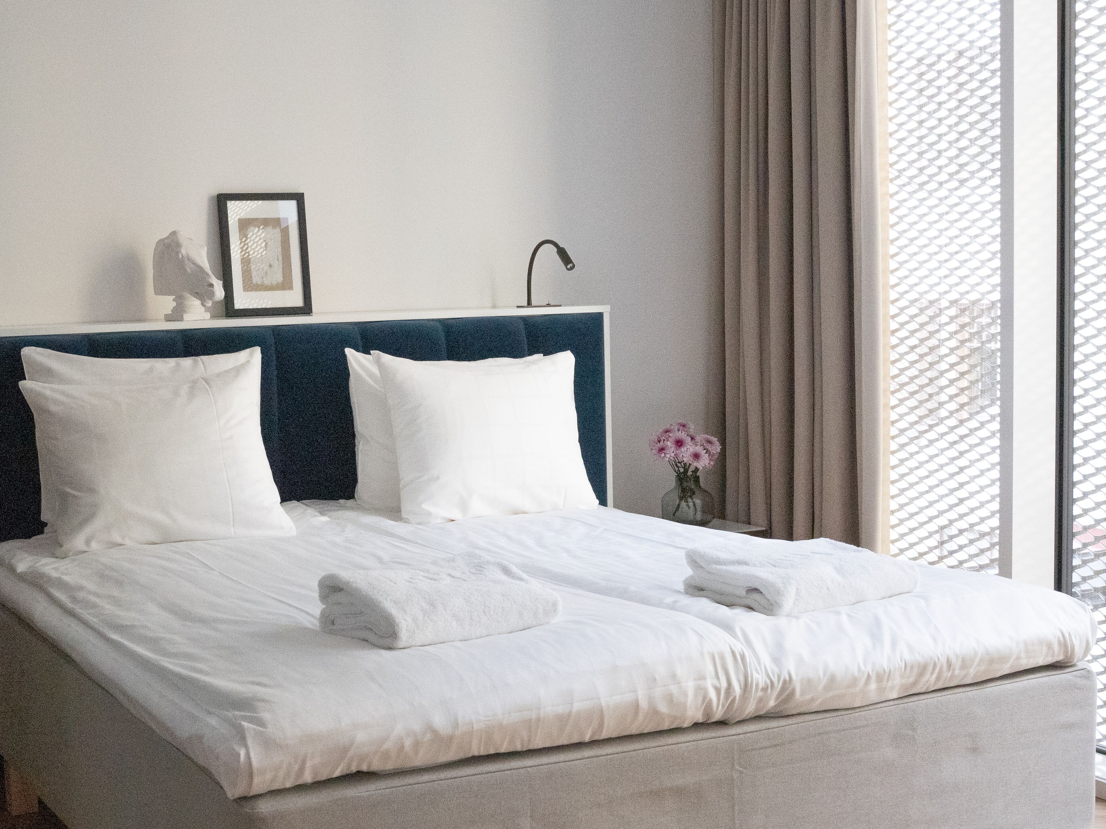 A neatly made bed with white linens and two pillows, a bedside table with a vase of flowers, and a window with a view of the outside.
