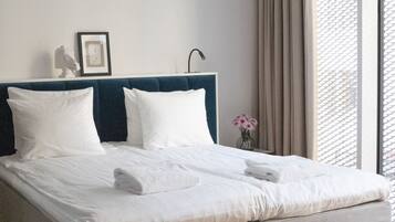 A neatly made bed with white linens and two pillows, a bedside table with a vase of flowers, and a window with a view of the outside.