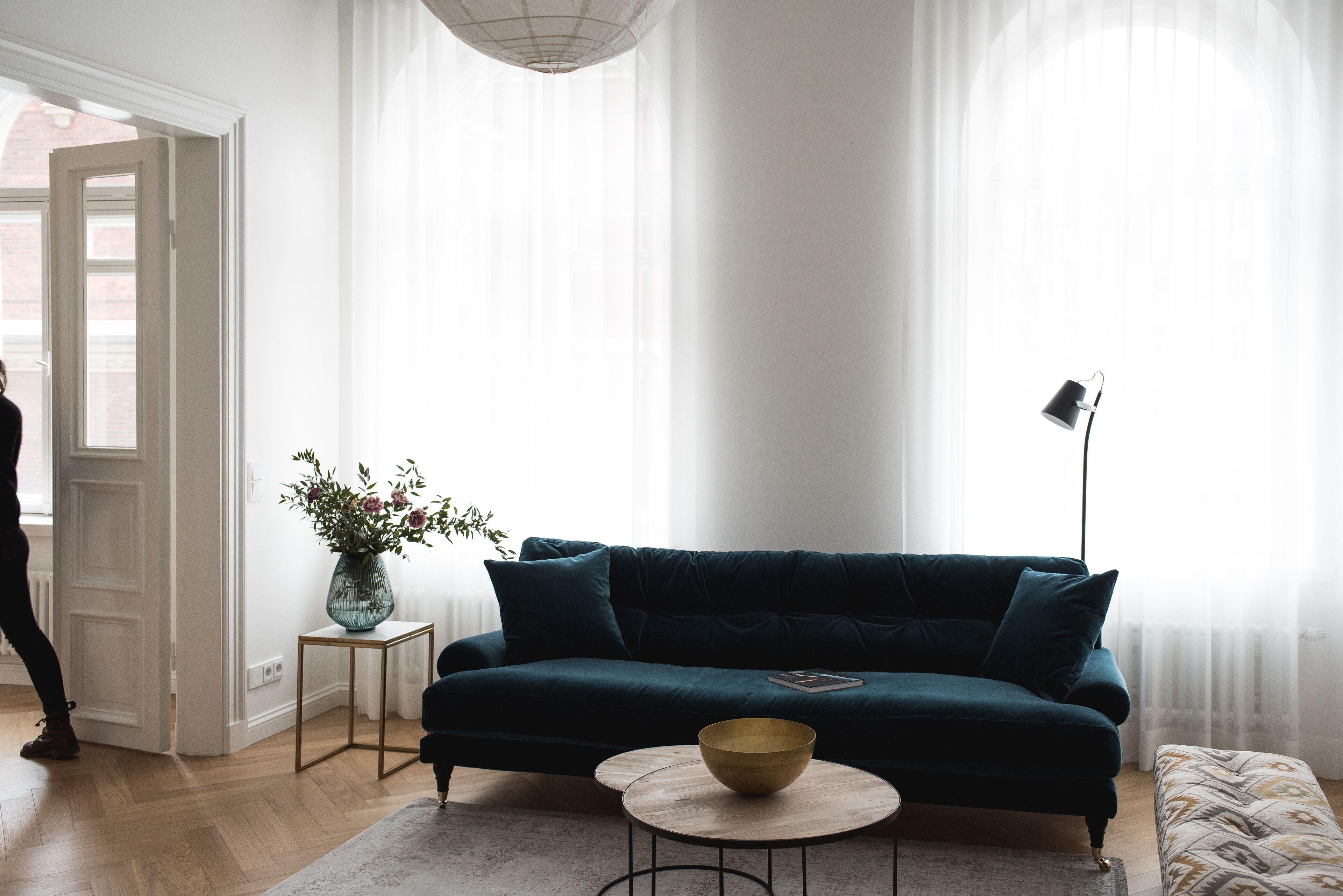 A living room with a dark green sofa, a small round table with a bowl, a floor lamp, and a vase with flowers on a side table.
