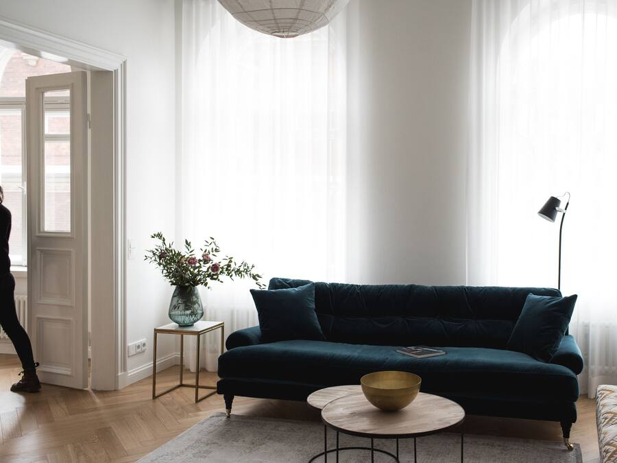 A living room with a dark green sofa, a small round table with a bowl, a floor lamp, and a vase with flowers on a side table.